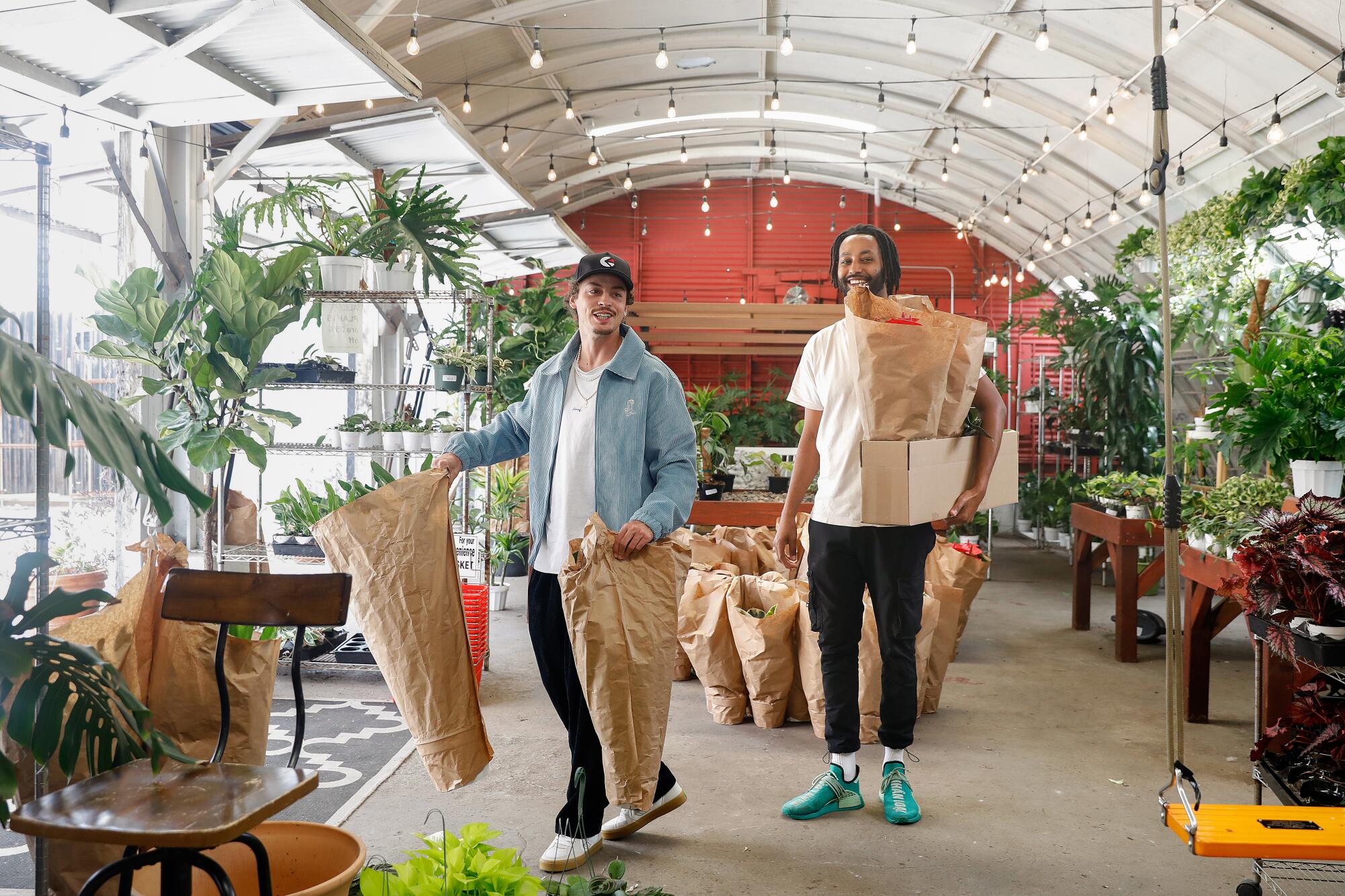 Two people carry plants wrapped in brown paper out of a greenhouse-like plant shop.