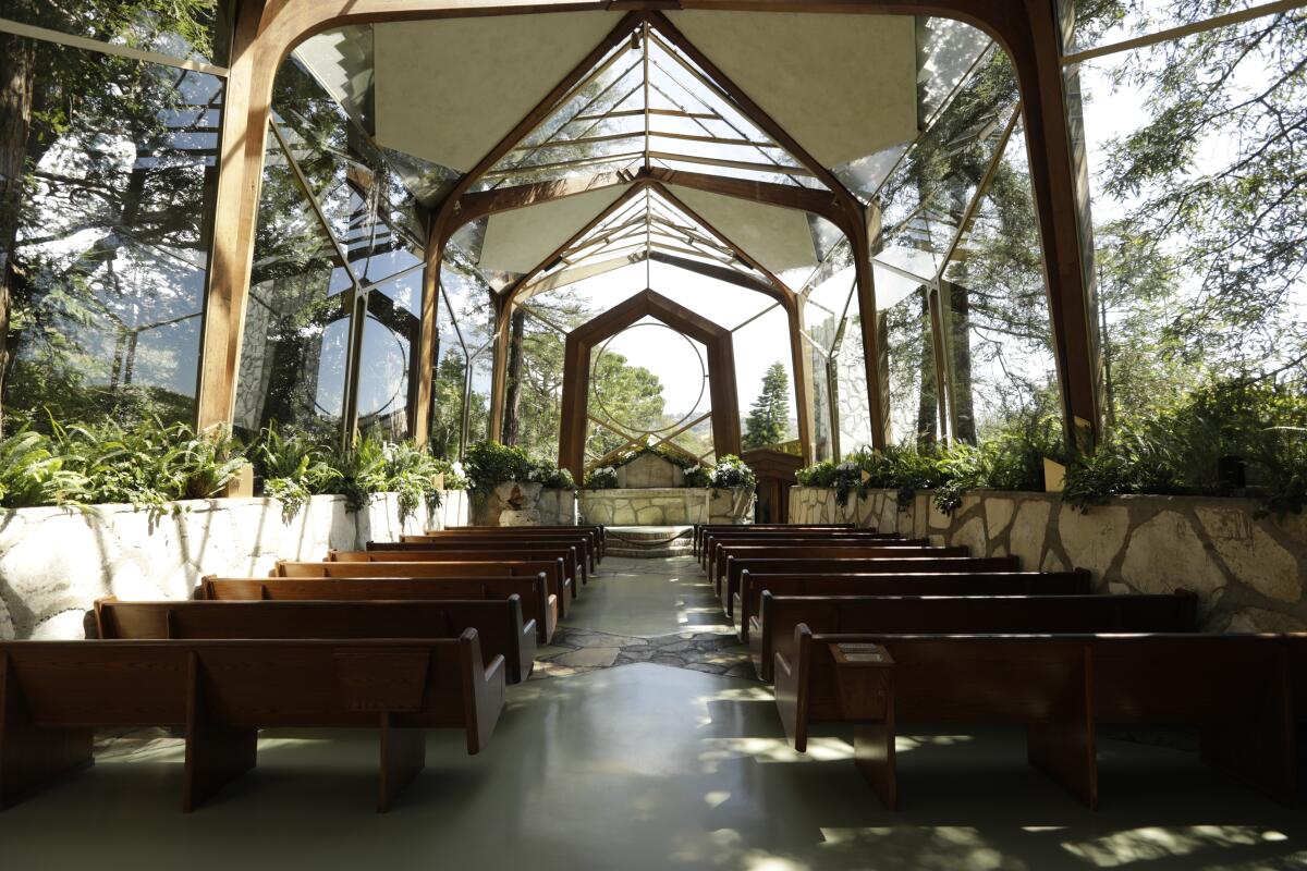 Trees are seen through the glass walls of a chapel. Inside, rows of pews are bordered by potted greenery.
