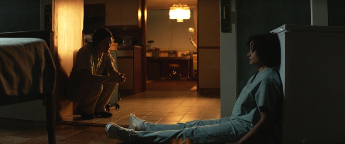 Two exhausted women sit on the floor of a dimly lit apartment.