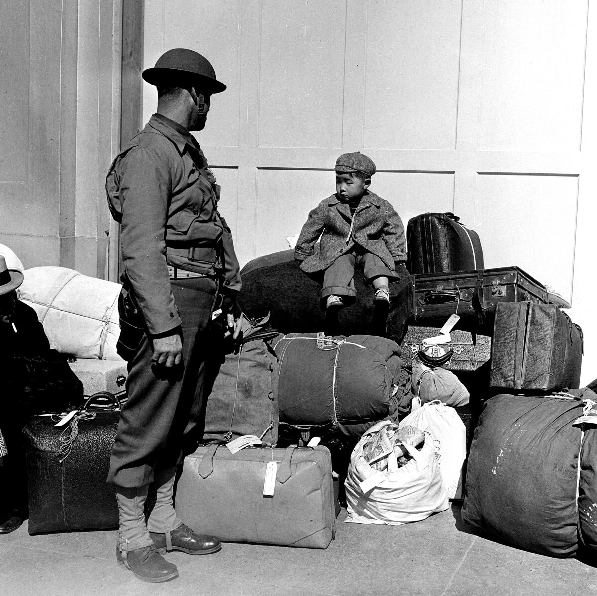 A boy sits on a pile of baggage as a military policeman watches him.
