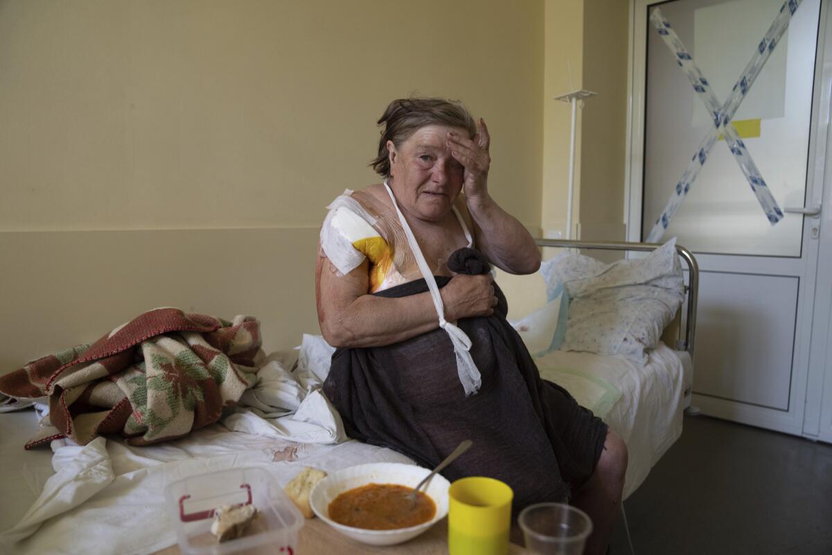 A woman sits on a bed, her shoulder bandaged, near a bowl of soup and yellow cup on a tray 