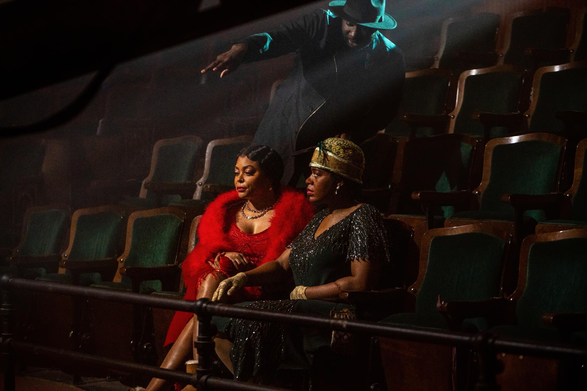 Two women in costume sit in a theater balcony as a man stands over them, pointing