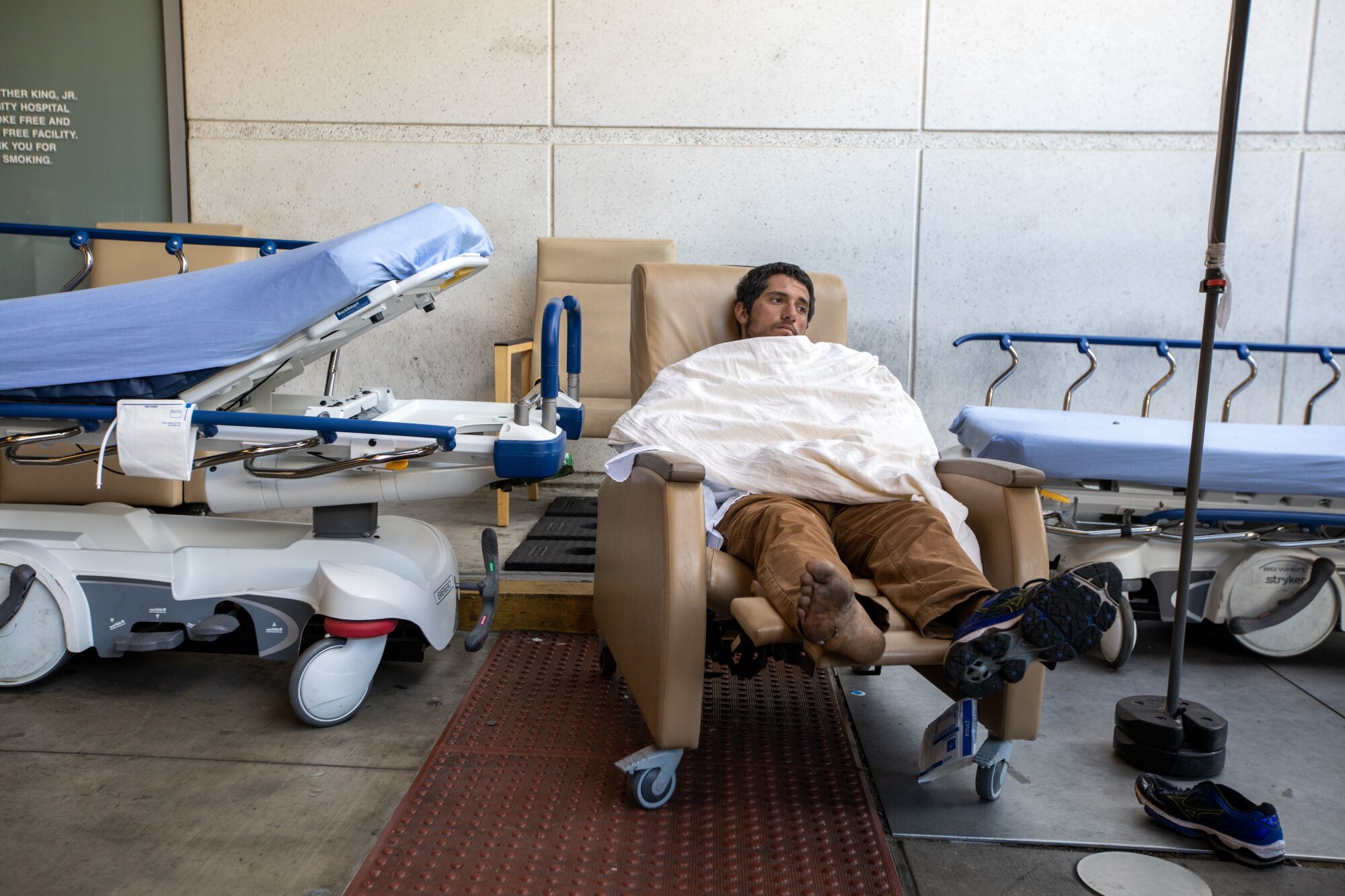 A man in one shoe sits under a blanket between empty hospital beds