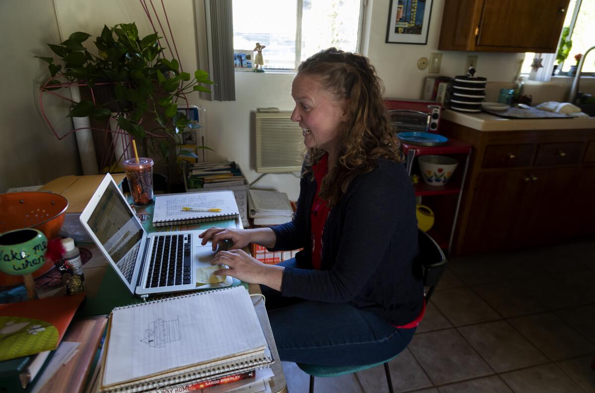 A seated woman works on a laptop on a table in a kitchen.