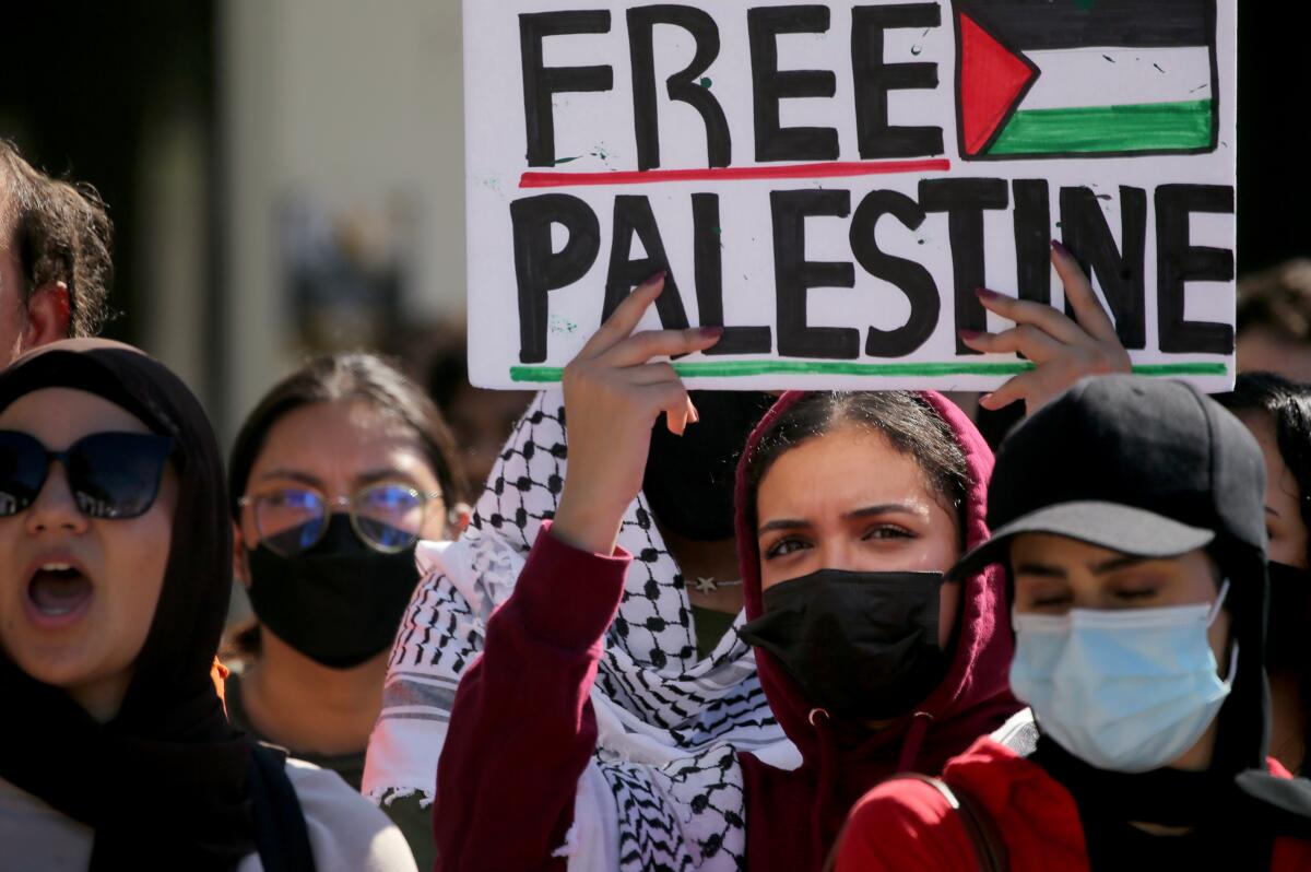 A woman in a black mask and red hoodie holds up a sign that reads "Free Palestine"