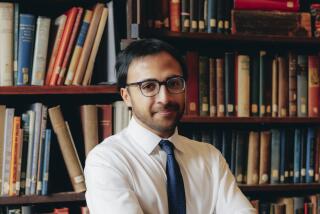 A man with glasses in a white dress shirt and blue tie stands in front of bookcases.