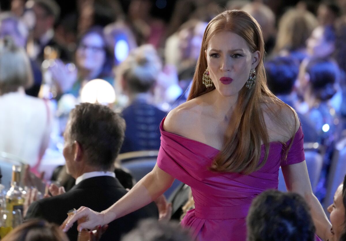 A woman with long red hair in a pink gown pursing her lips and walking through a crowd of people.