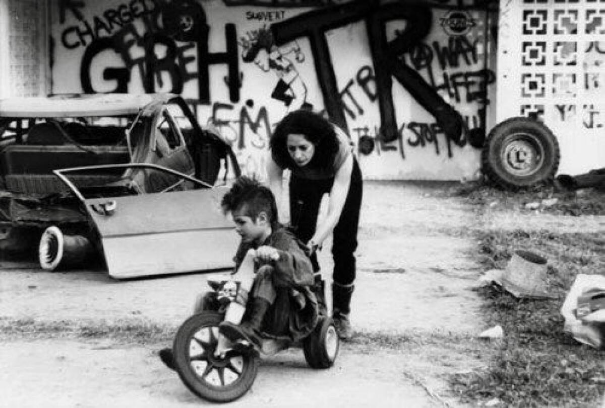 A woman pushes a young boy with spiked hair on a tricycle.