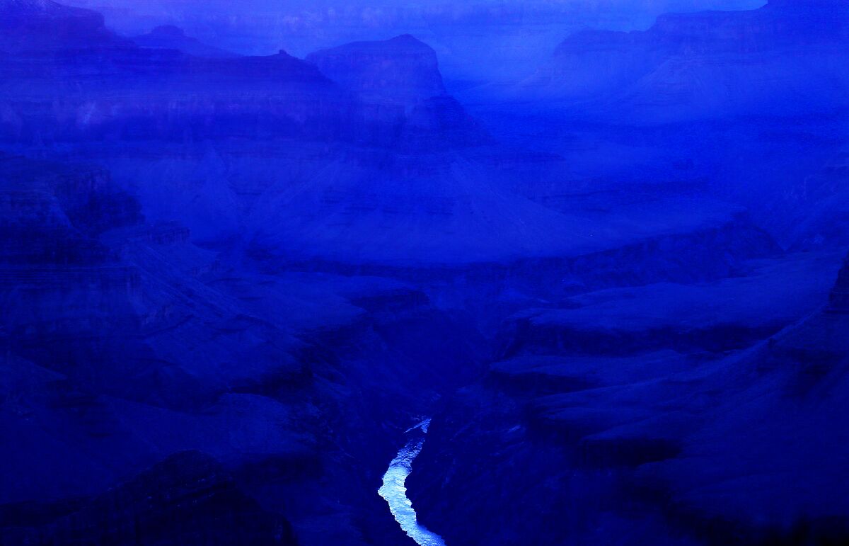 Mountains and mesas near dark are in shades of indigo. A river trickles in the foreground, with a mirror-like surface.