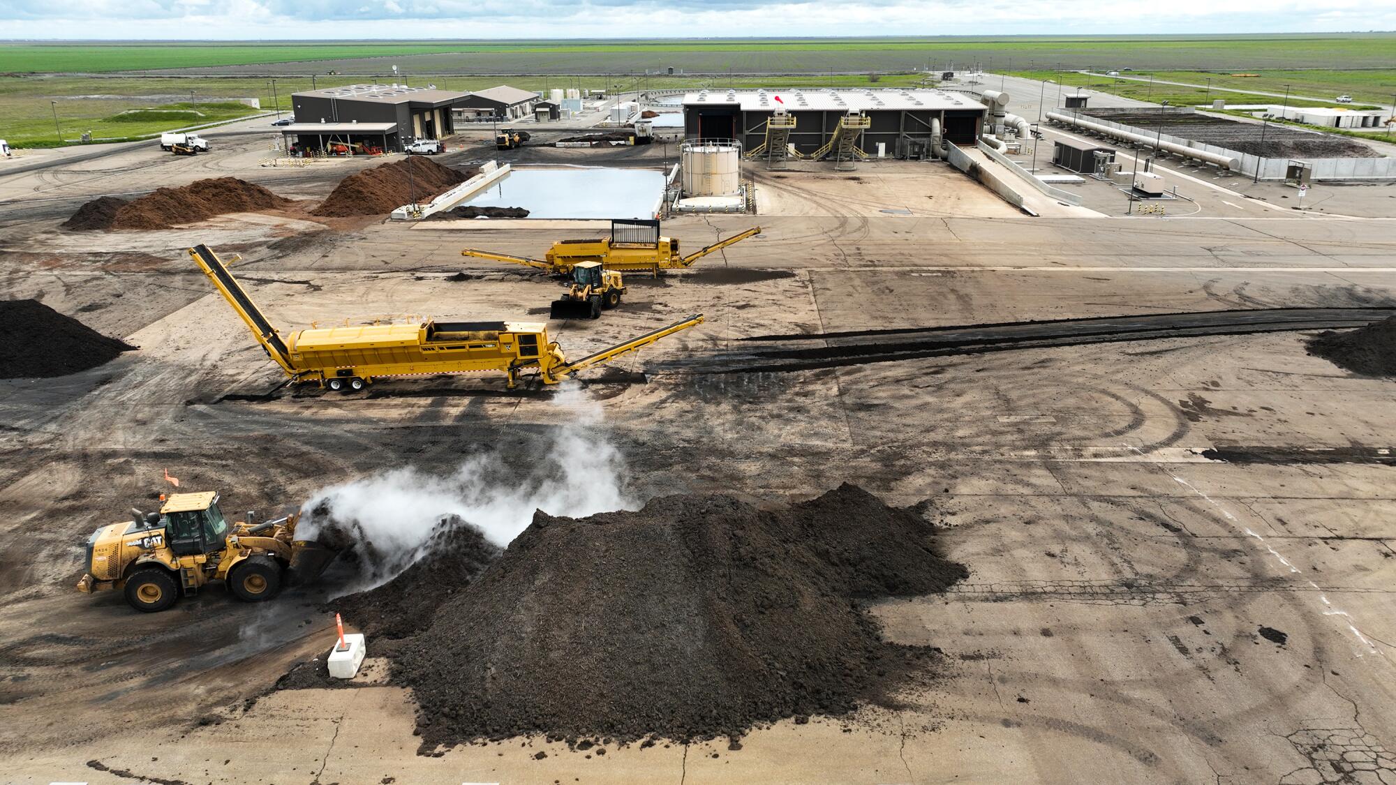 A piece of heavy machinery pushes a pile of dark compost as vapor rises from the material.