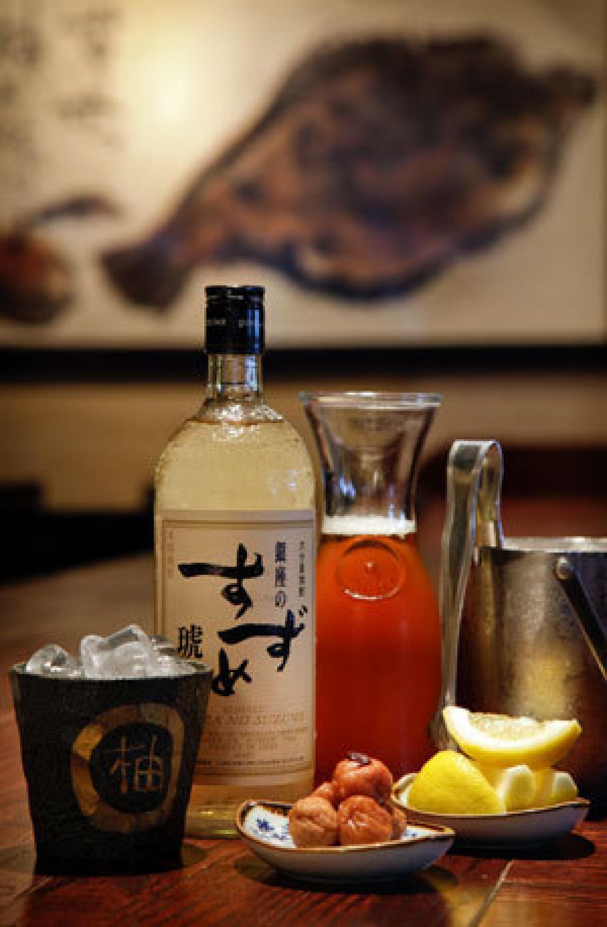 A Ginza No Suzume Kohaku shochu bottle and a carafe of oolong tea, pickled plums and lemon wedges on the counter at Yuzu Izakaya in Torrance.