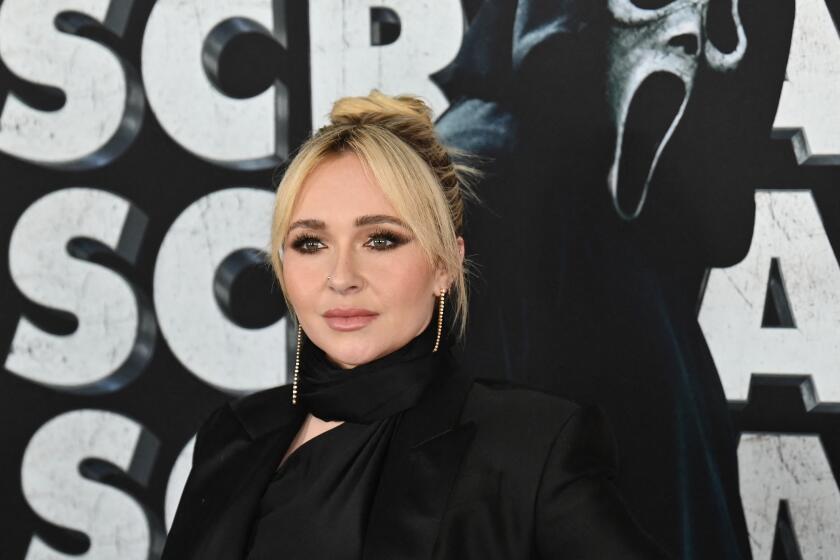 Woman, Hayden Panettiere standing on red carpet dressed in all black, flanked by poster of Ghostface from Scream 6 