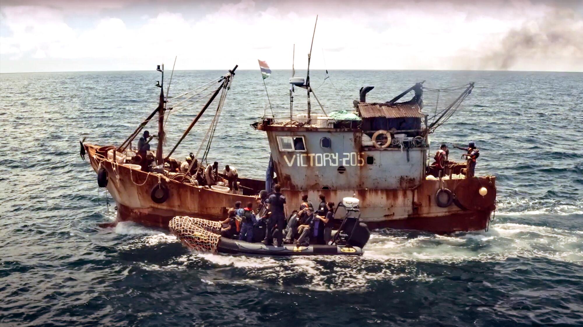 A rust-covered fishing boat with the name Victory and a dinghy carrying people next to it, on the ocean
