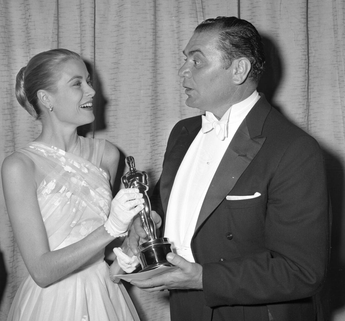 A man in white tie and tails is handed a small statuette by a woman in a strapless gown and short white gloves.