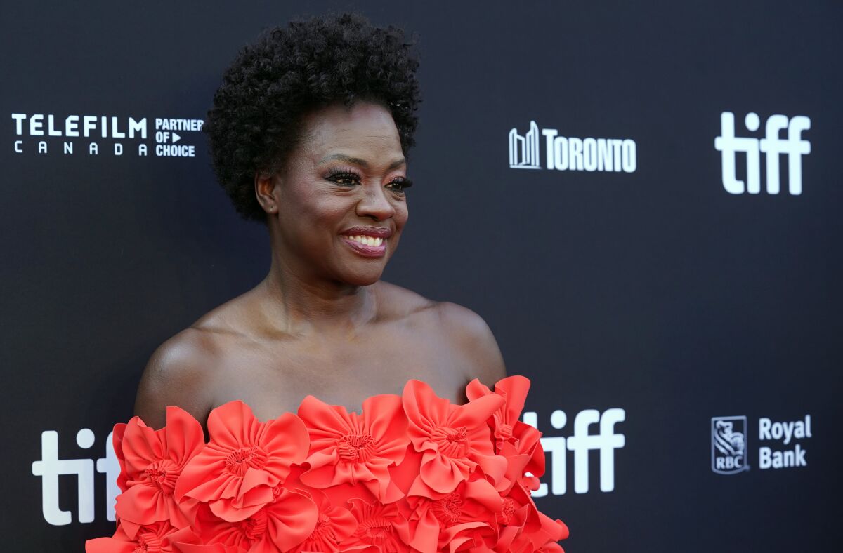 A woman wearing an orange dress with fabric flowers smiles while standing in front of a backdrop.