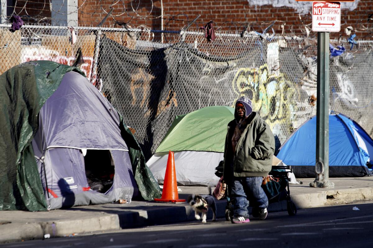 A man walks with his dog along a sidewalk with tents and a fence with graffiti 