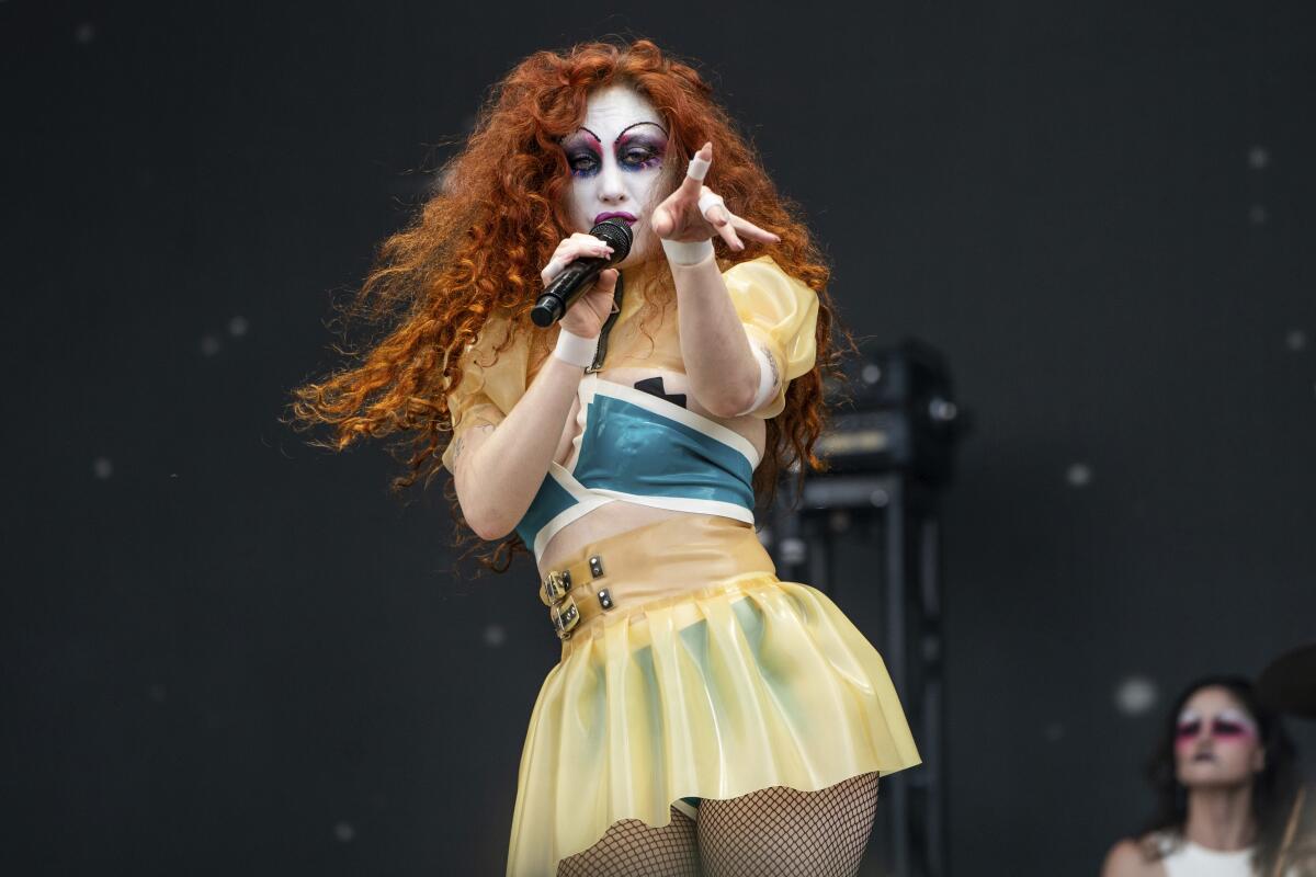 Chappell Roan pointing while singing into a microphone, wearing a light-colored dress and white face paint. 