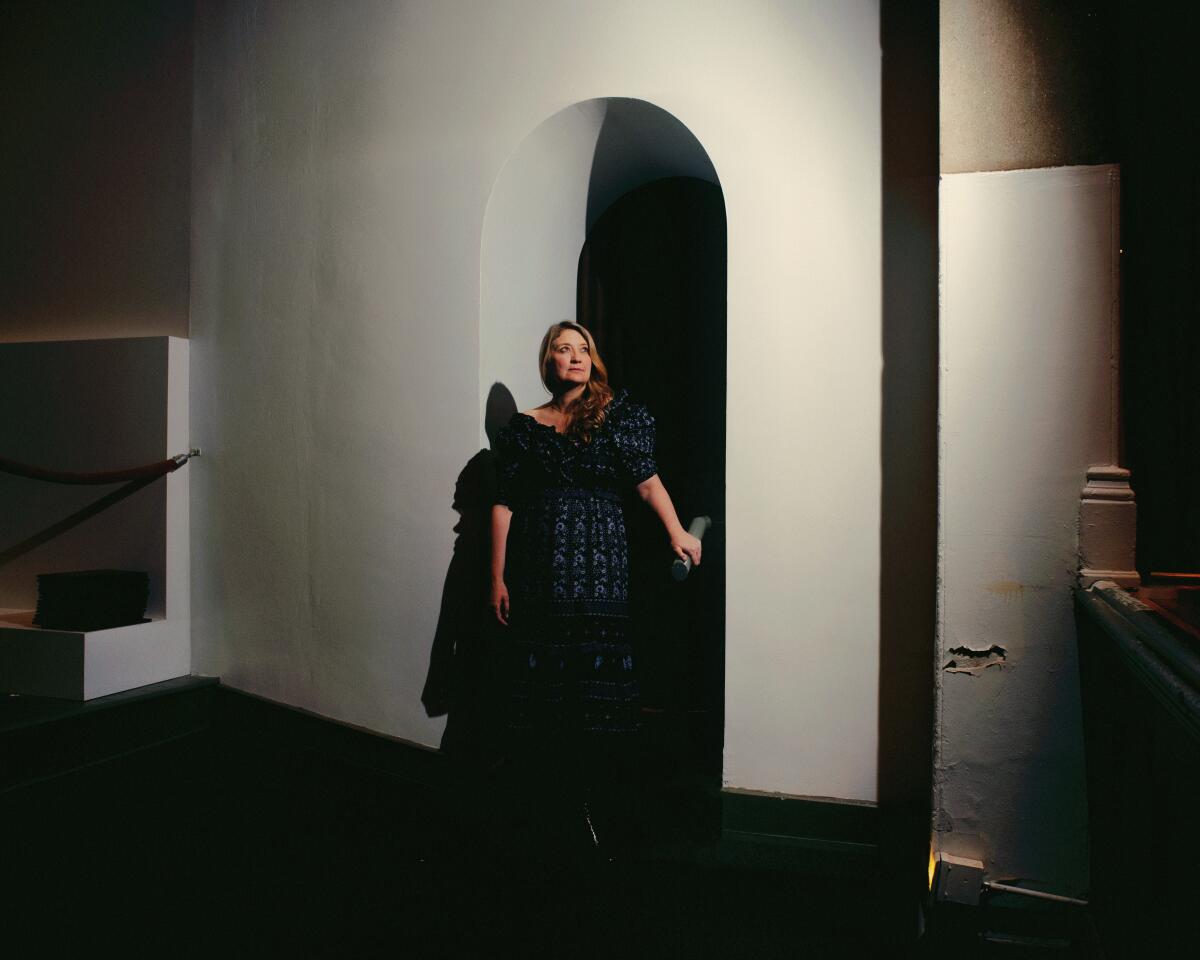 Heidi Schreck, wearing a flowered dress and bathed in dramatic light, stands in an arched doorway.