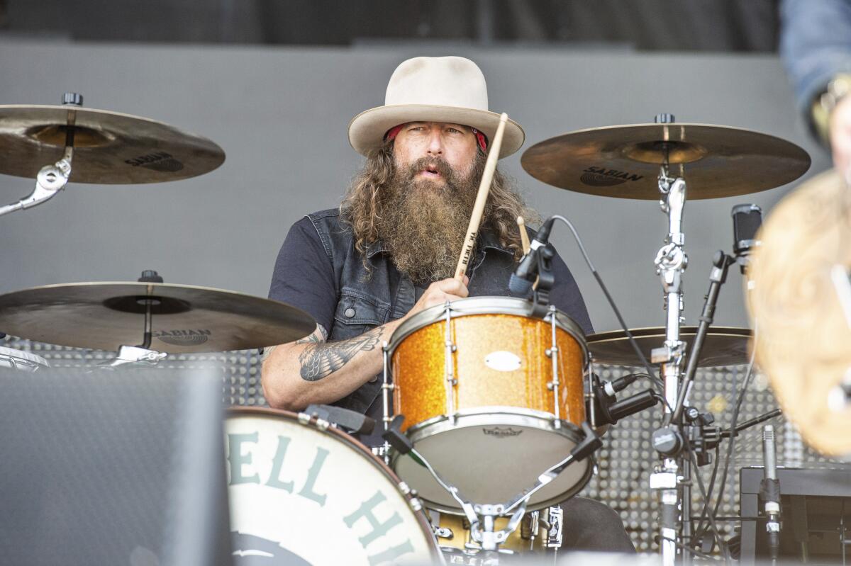A man in a cream, wide-brimmed hat with a beard and tattoos on his forearm sitting at and playing a drumkit on a stage