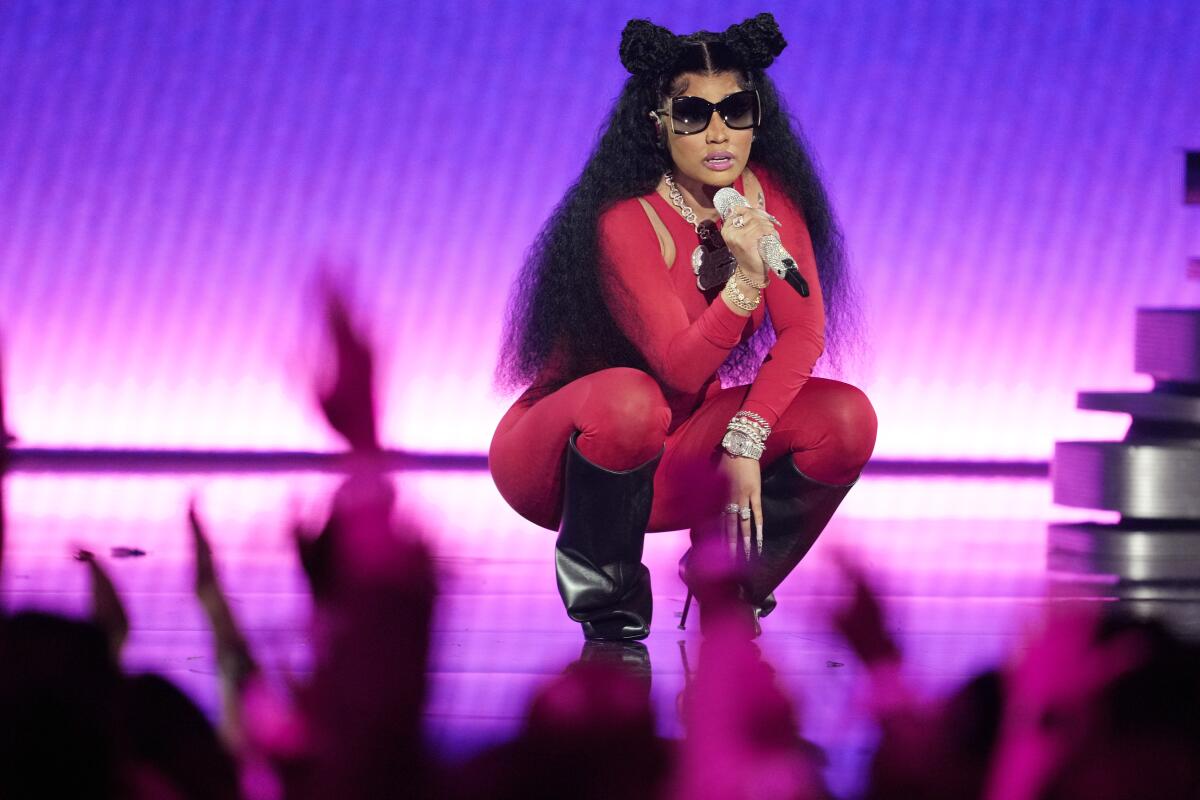 Nicki Minaj holds a mic to her mouth while squatting onstage in a red bodysuit, black boots and dark sunglasses