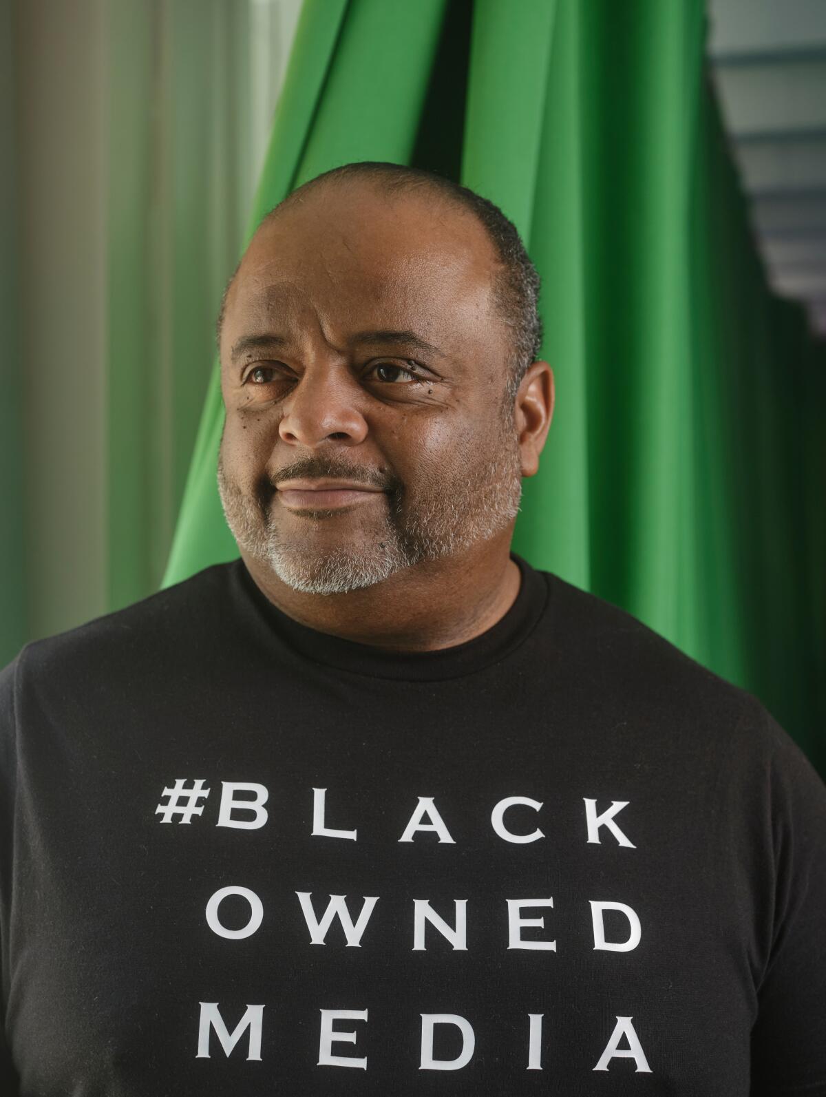 A man stands in a black T-shirt stands in front of green screen fabric.