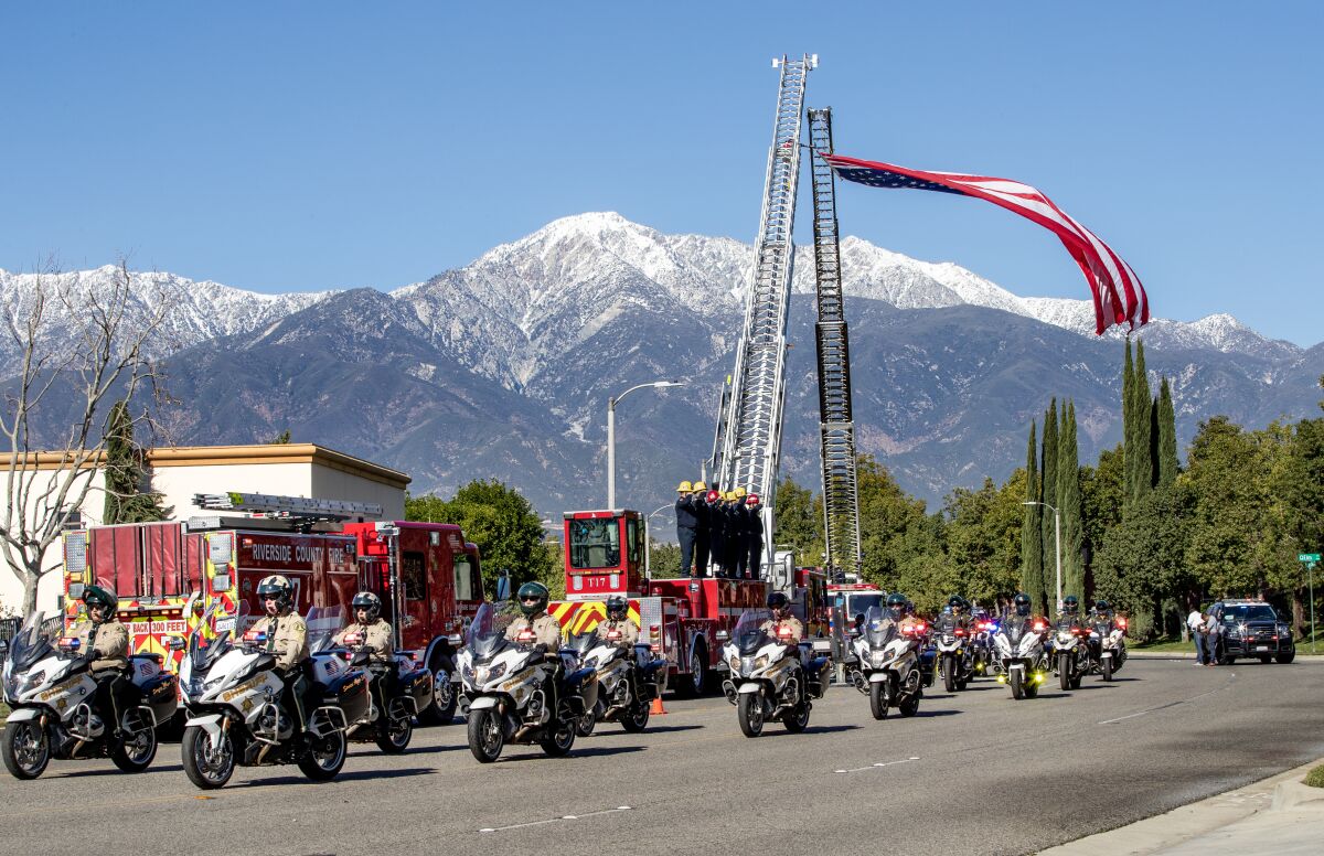Deputies on motorcycles pass firetrucks as an American flag hangs between the trucks' raised ladders