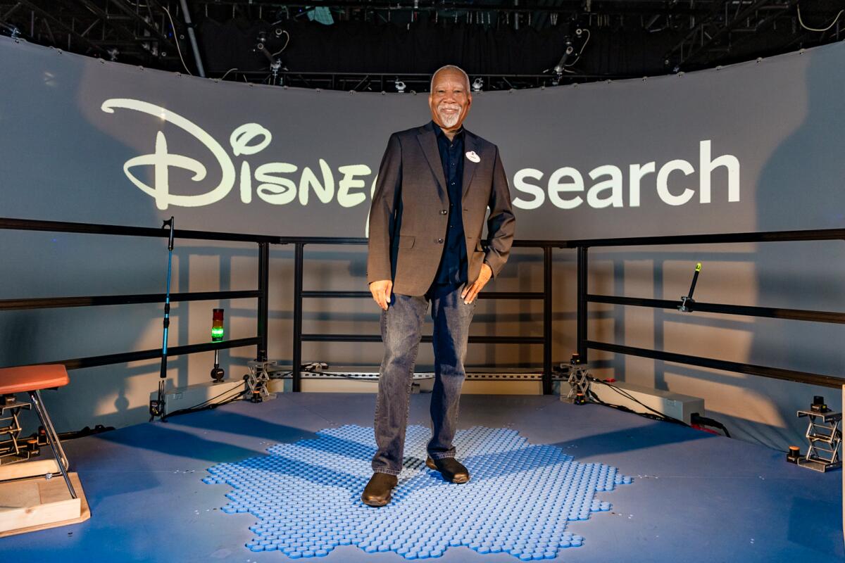 A goateed man in a blazer over jeans stands on blue tiles, smiling, in front of a sign that says Disney Research