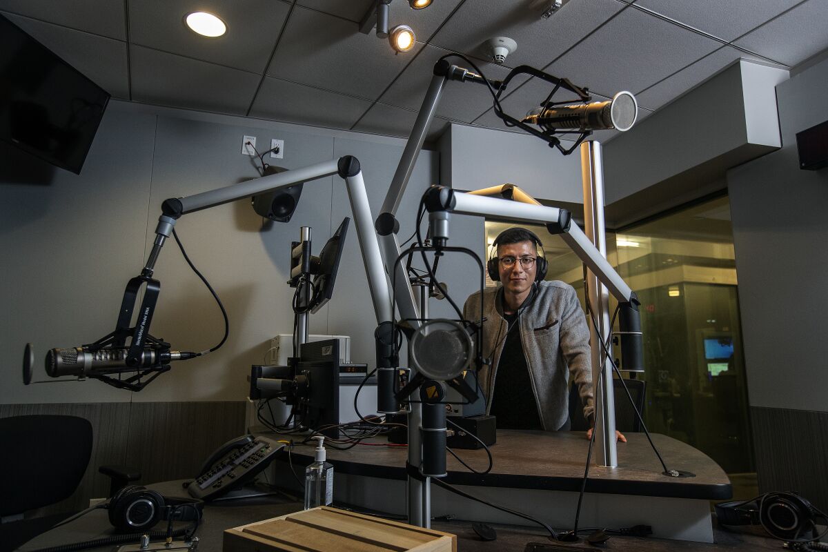 A man wearing headphones stands inside an audio recording studio, which features a desk and several microphones.