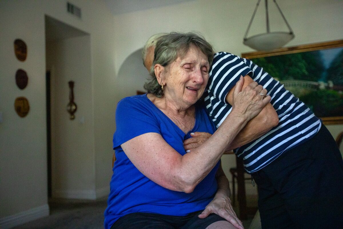 A woman sits facing the camera with her eyes closed and crying while being embraced by another person. 