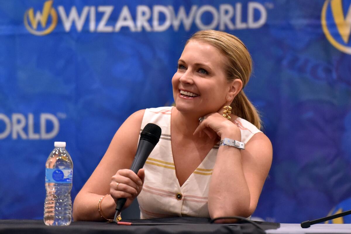 Melissa Joan Hart smiles while leaning on a table and holding a microphone