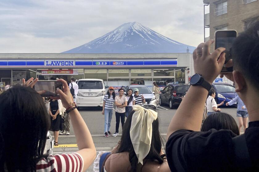 Visitors take a photo in front of a convenient store at Fujikawaguchiko town, Yamanashi prefecture, Japan, with a backdrop of Mr. Fuji on April 28, 2024. The town of Fujikawaguchiko, known for a number of popular photo spots for Japan's trademark of Mt. Fuji, on Tuesday, April 30, 2024 began to set up a huge black screen on a stretch of sidewalk to block view of the mountain in a neighborhood hit by a latest case of overtourism in Japan. (Kyodo News via AP)