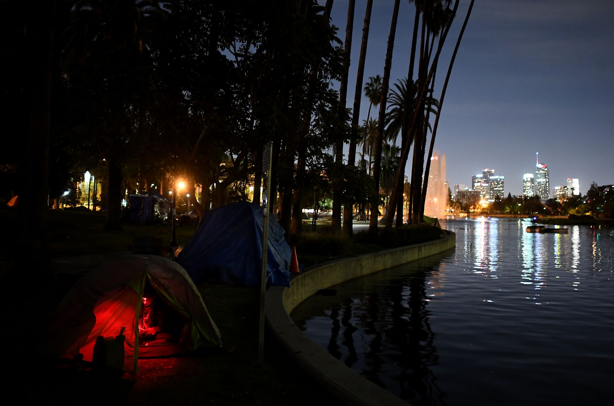A glow is visible inside a tent along a lake after dark, with the city skyline in the background.