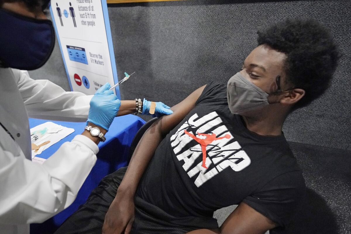 A health worker in protective gear and holding a syringe bends over a seated young man with a face tattoo.