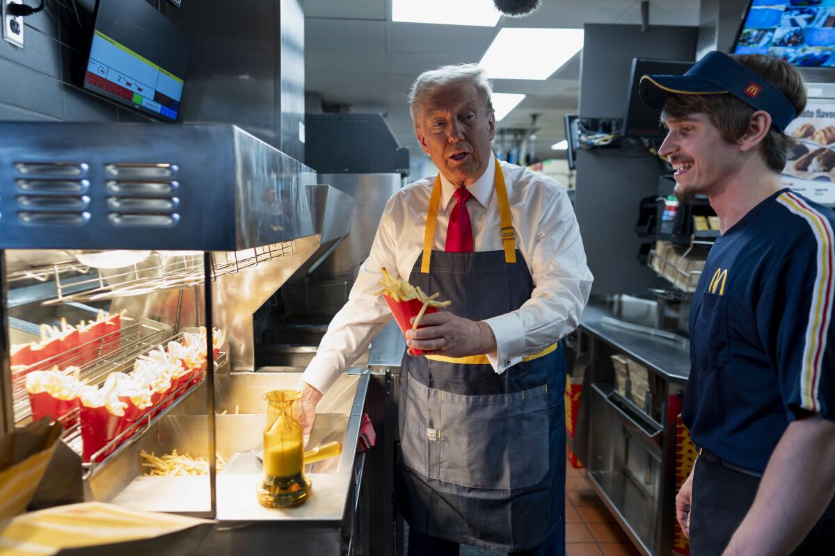 A man with blond hair, in white shirt, red tie and dark apron, holds a red carton of fries as another man smiles next to him