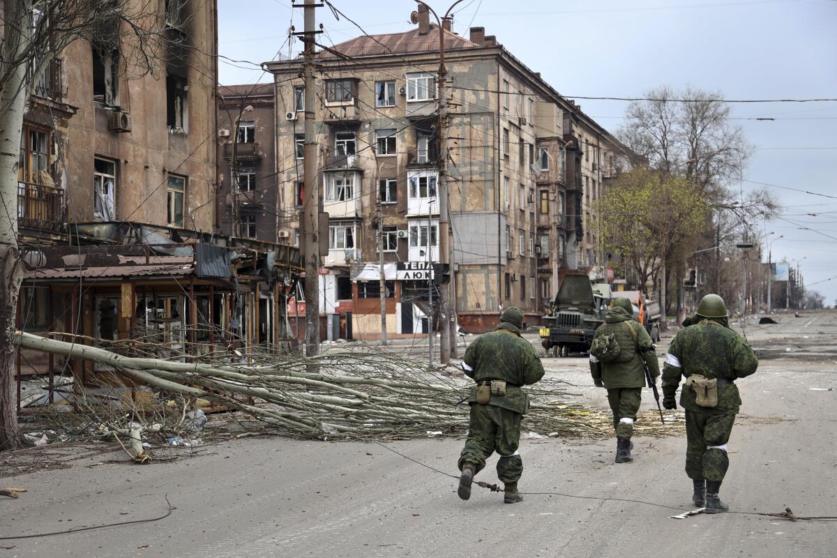 People in combat gear walk past damaged buildings and a fallen tree.