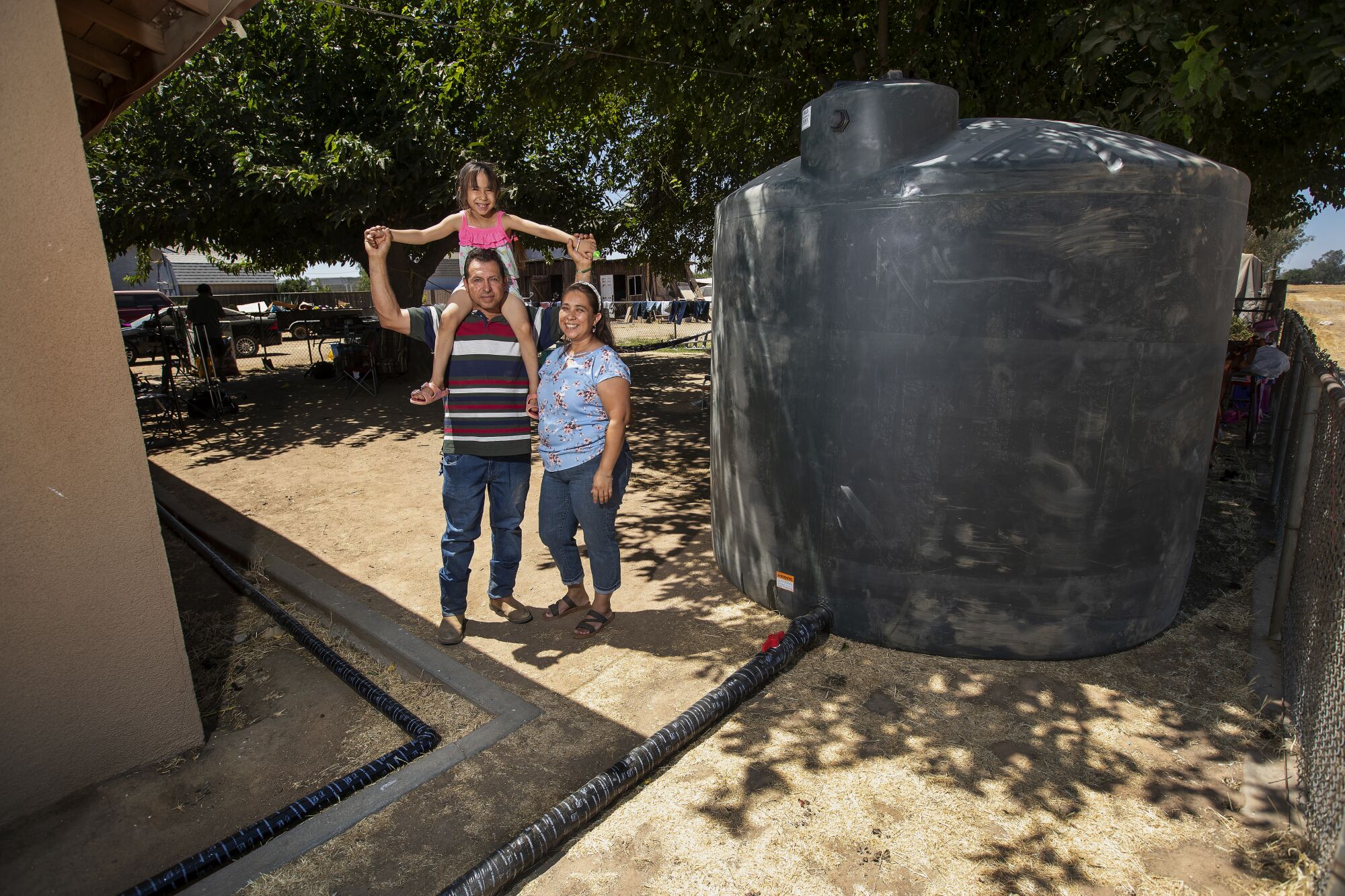 A girl sits on her father's shoulders and poses with him and her mother next to a large water tank