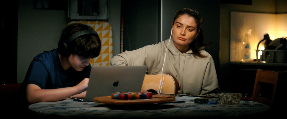 A teen boy looks at his laptop as a woman wearing earbuds plays a guitar in "Flora and Son." 