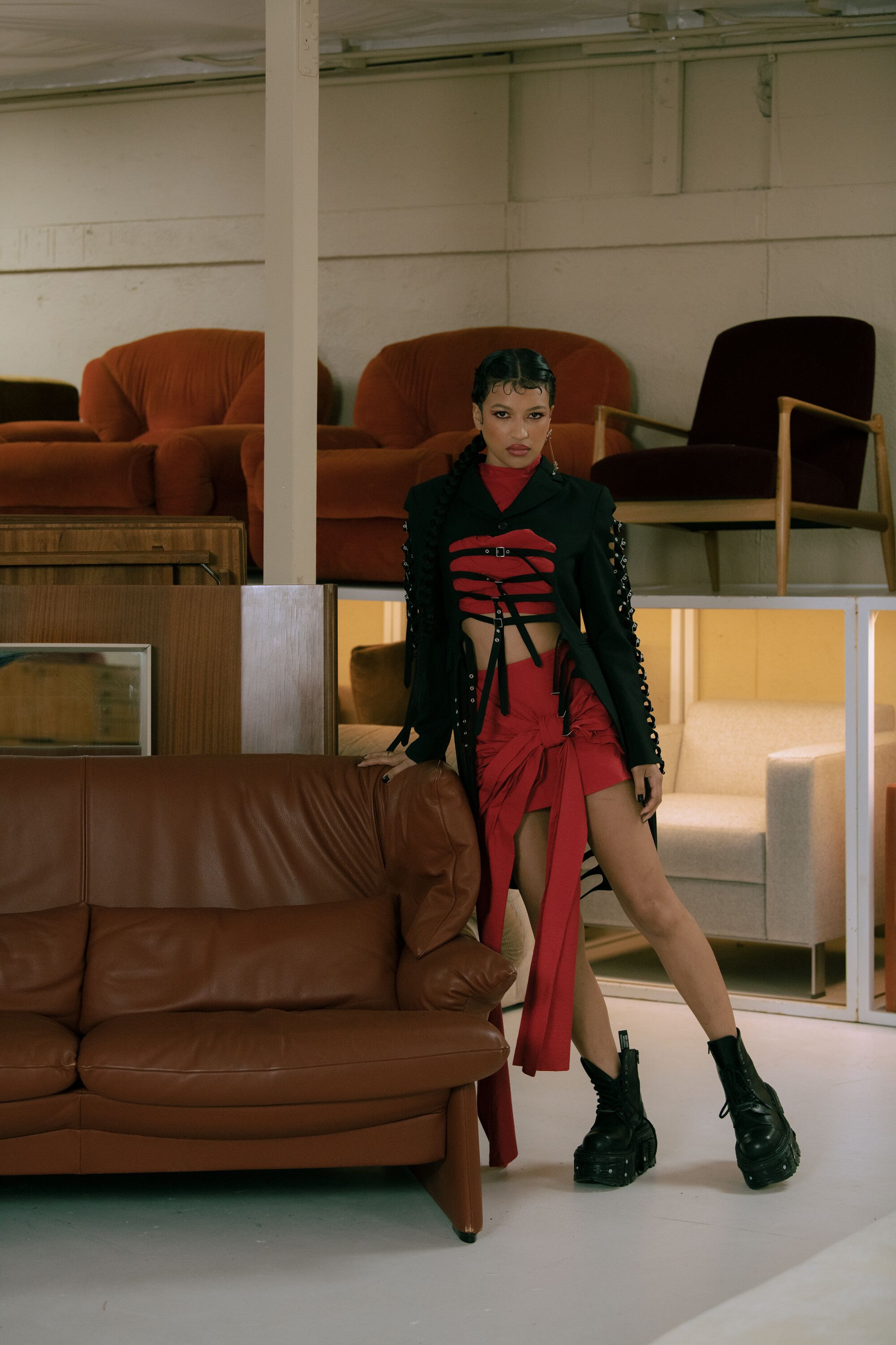 A model wearing a red and black dress leans against a brown leather couch.