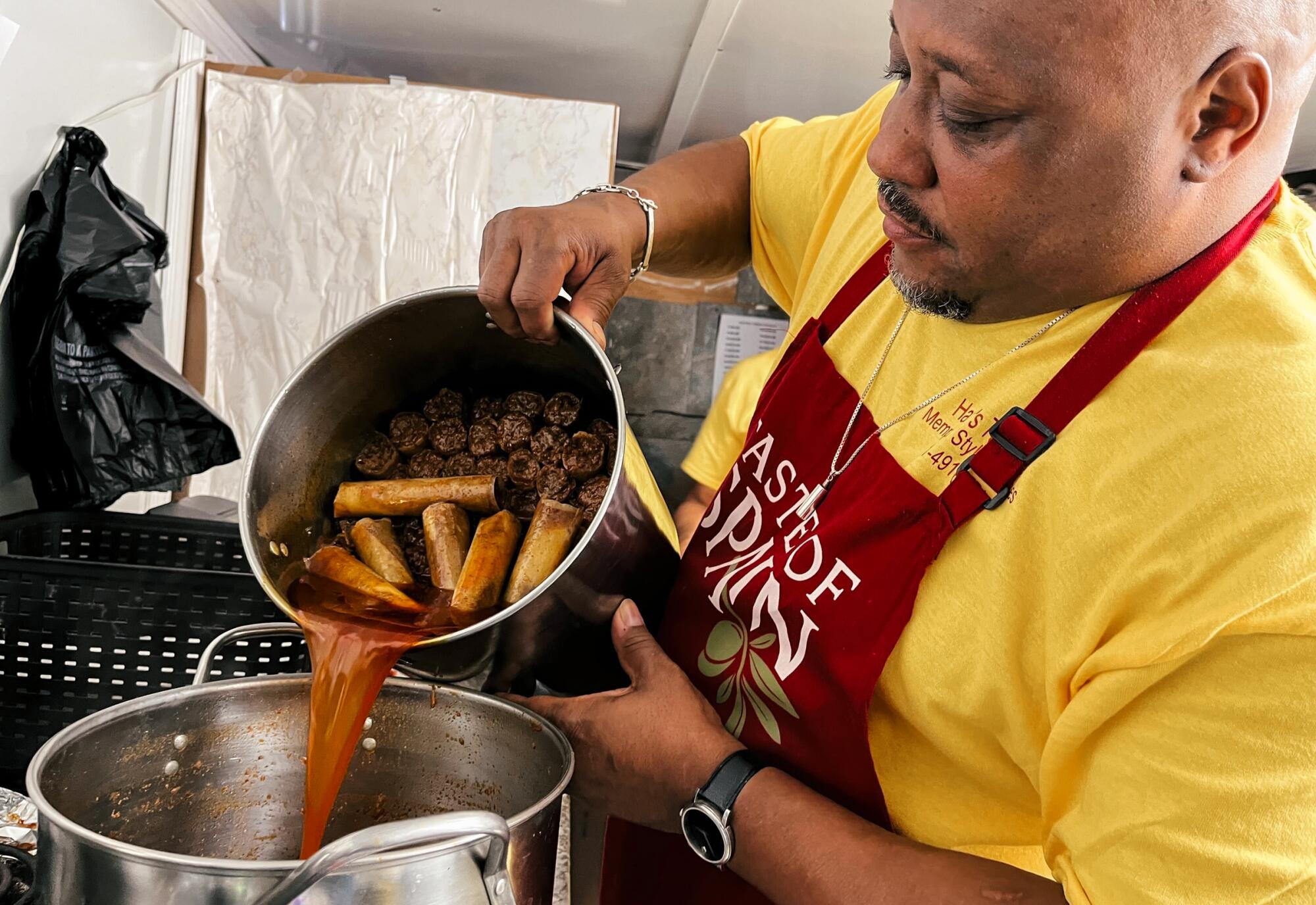 A man in a yellow T-shirt and red apron pours tamales and their sauce into a large pot.