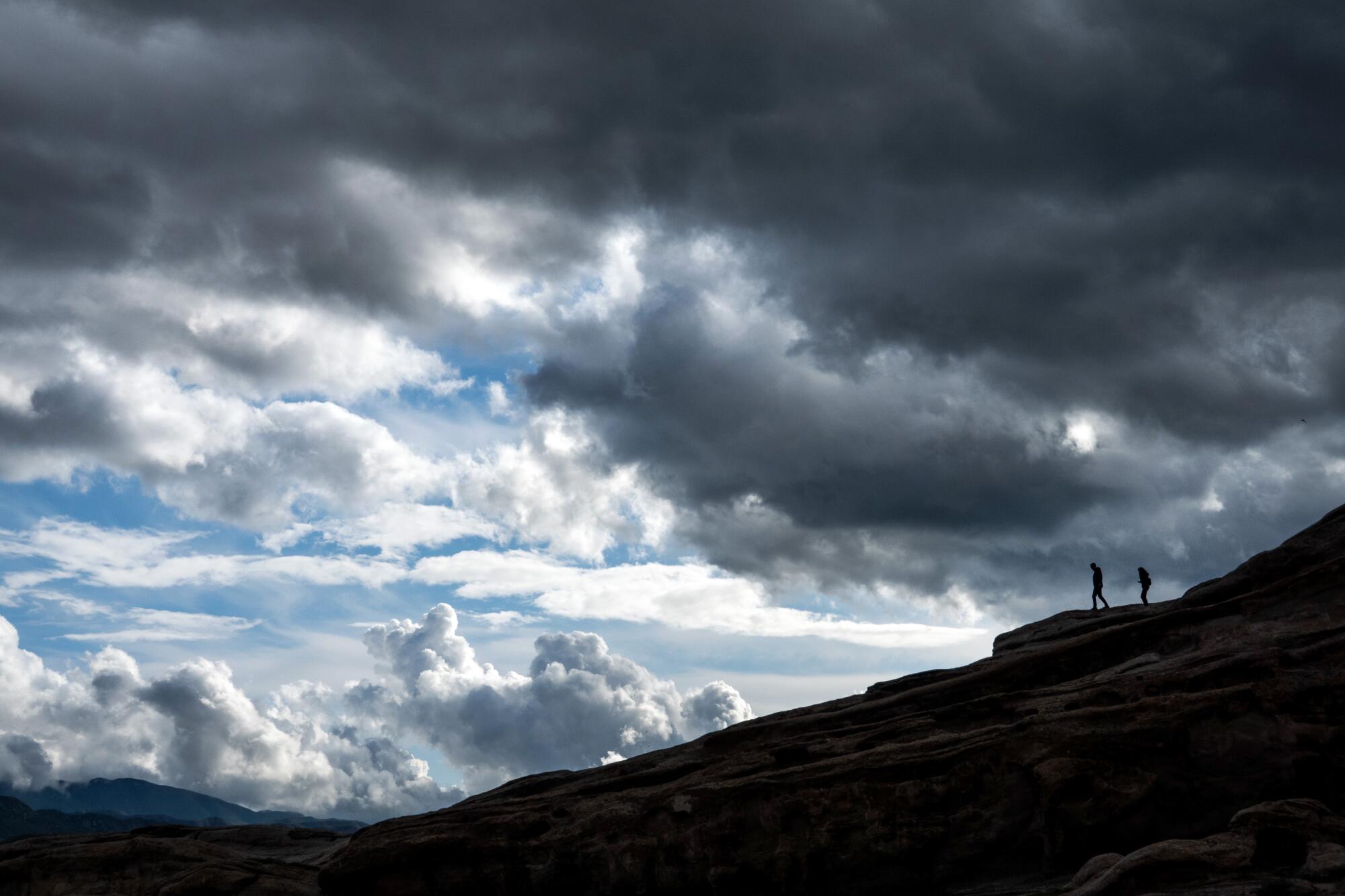 Two small figures standing on dark rocky terrain are silhouetted against a sky of heavy and fluffy clouds.