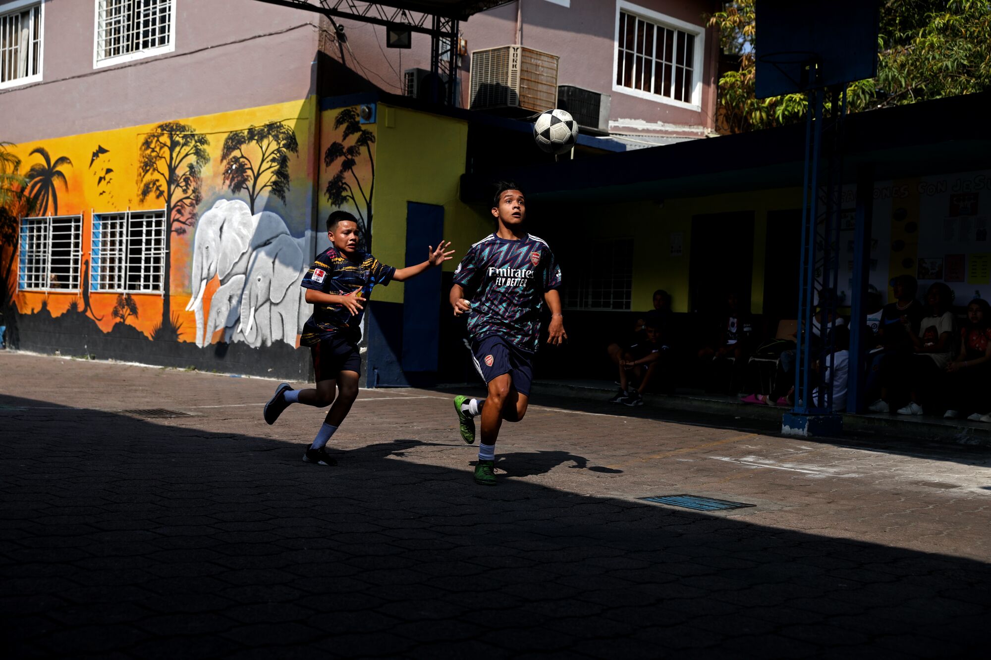 Two teens run down a street in front of a building with a mural in orange shades and white