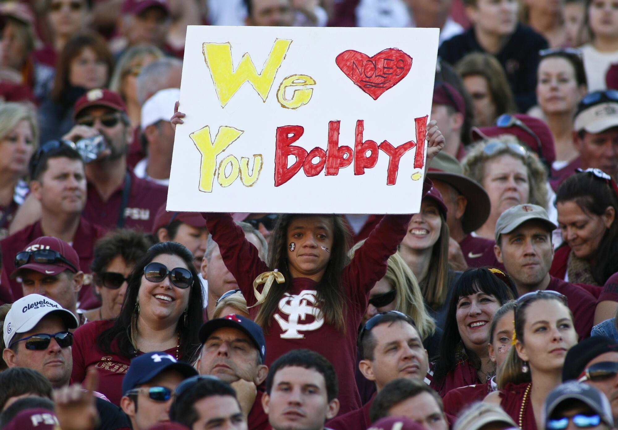In a crowd in the bleachers, a woman stands, holding a handwritten sign that says "We [heart] you Bobby."