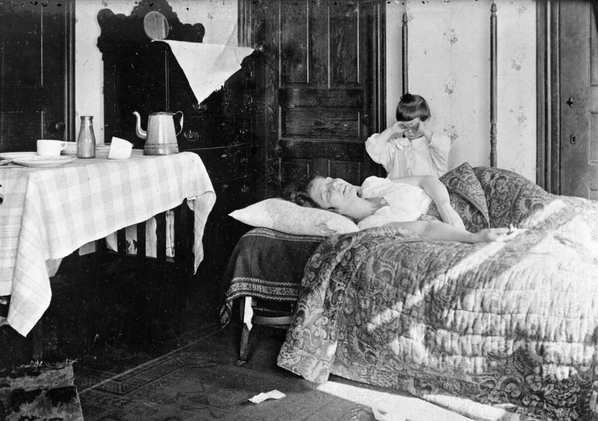 A girl stands next to her sister lying in bed in November 1918. 