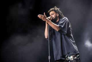 J. Cole in a dark shirt holding his left hand out in front of him while speaking into a microphone on a dark stage