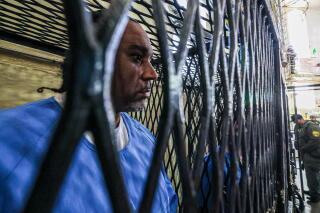 A man, in blue prison clothes, is seen through the enclosure of his cell