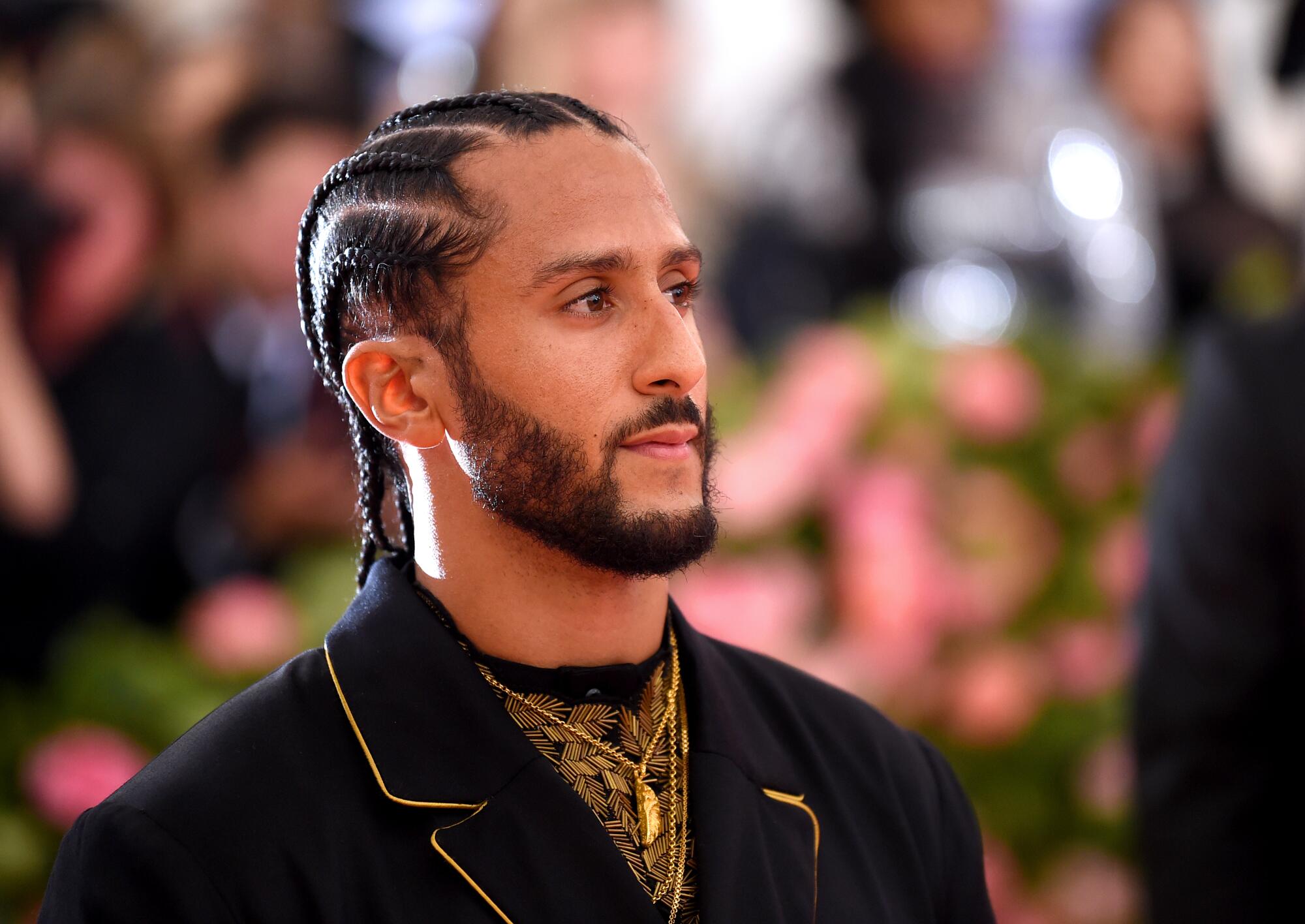 A man with braided hair and a beard posing in a black and gold suit