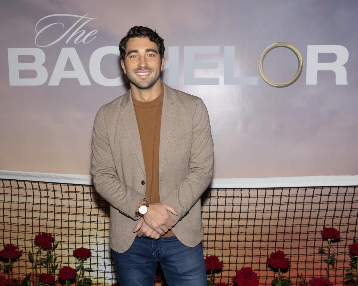 Joey Graziadei smiles in front of a Bachelor TV show poster
