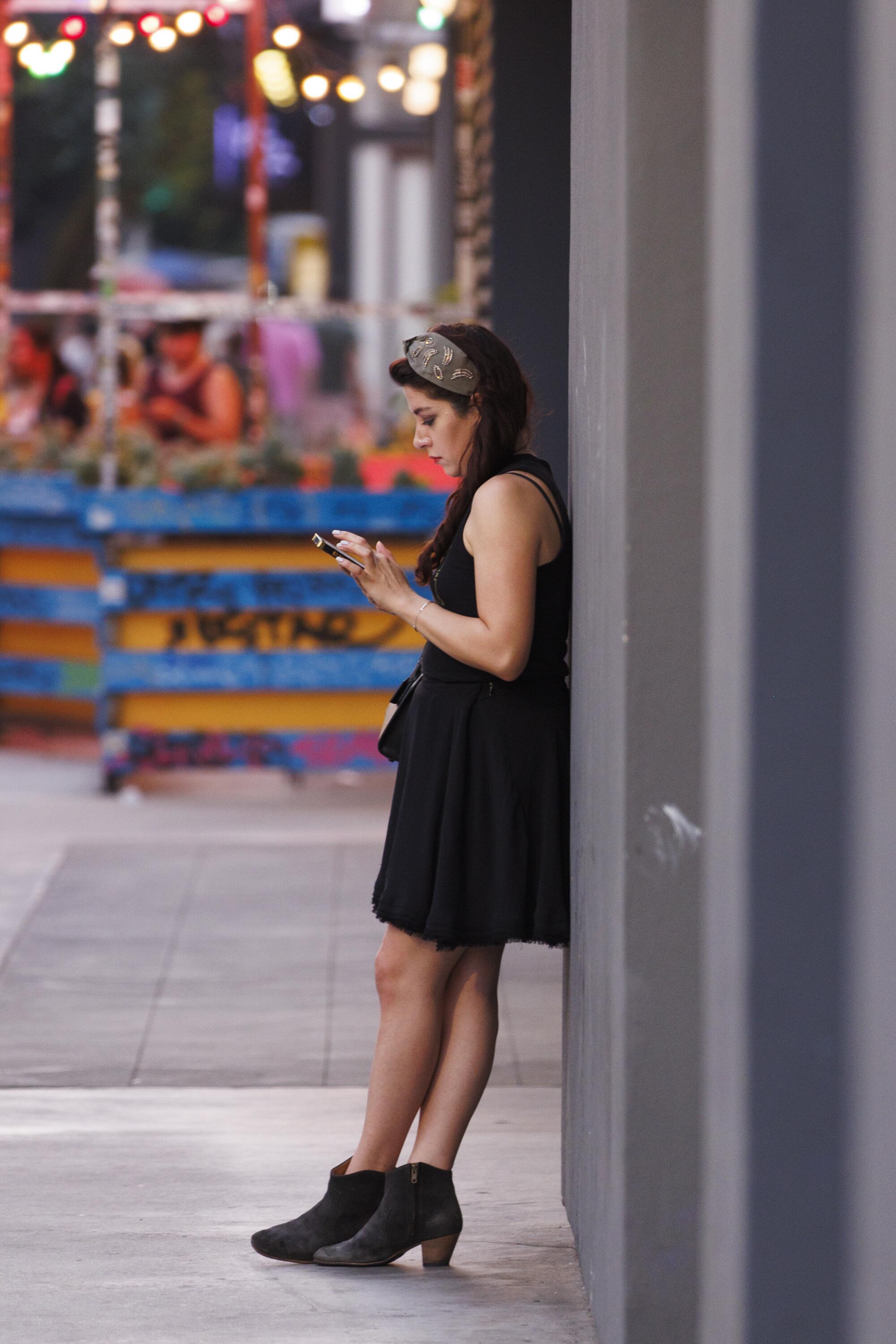 A woman leans with her feet crossed and back against a wall while looking at her phone