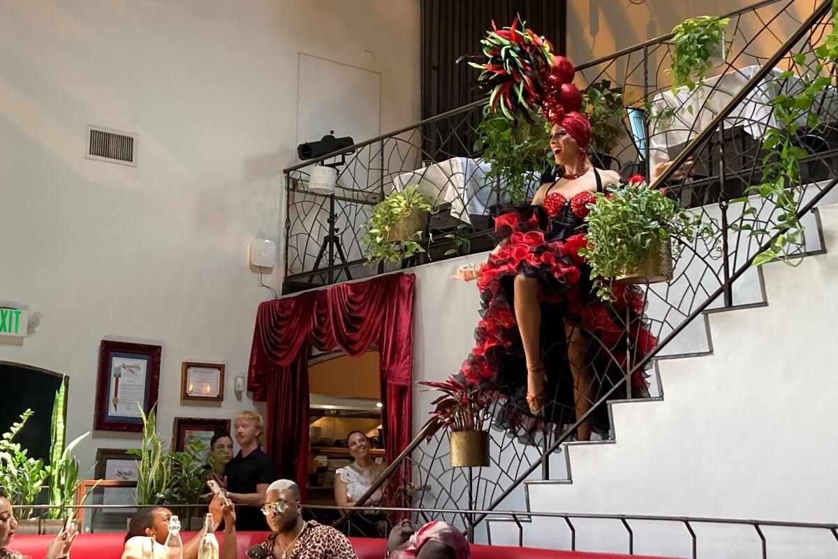 A drag queen in red velvet looks glamorous on a wrought iron stair bannister, with diners seated below.