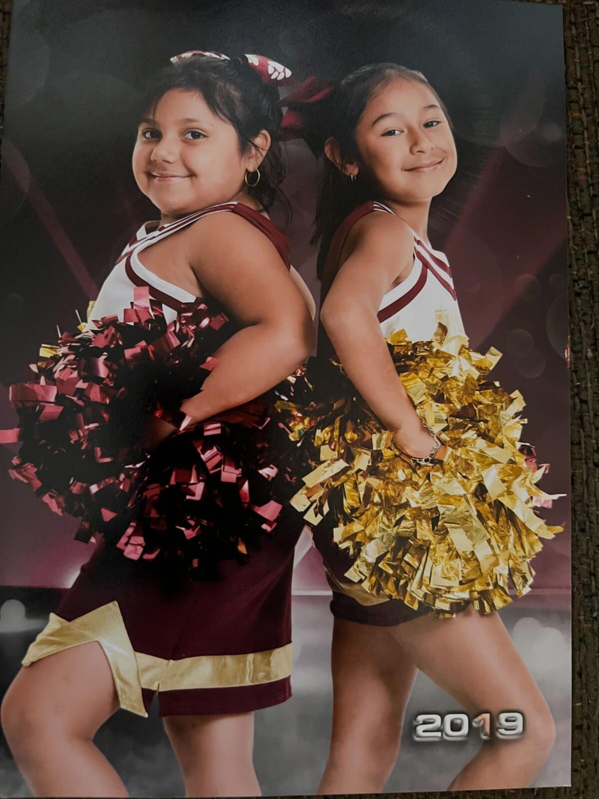 Two little girls in cheerleading uniforms holding pompoms and standing back-to-back